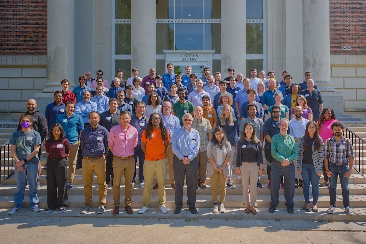 A group of about 60 people stand on the steps of an academic buiding to pose for a photo