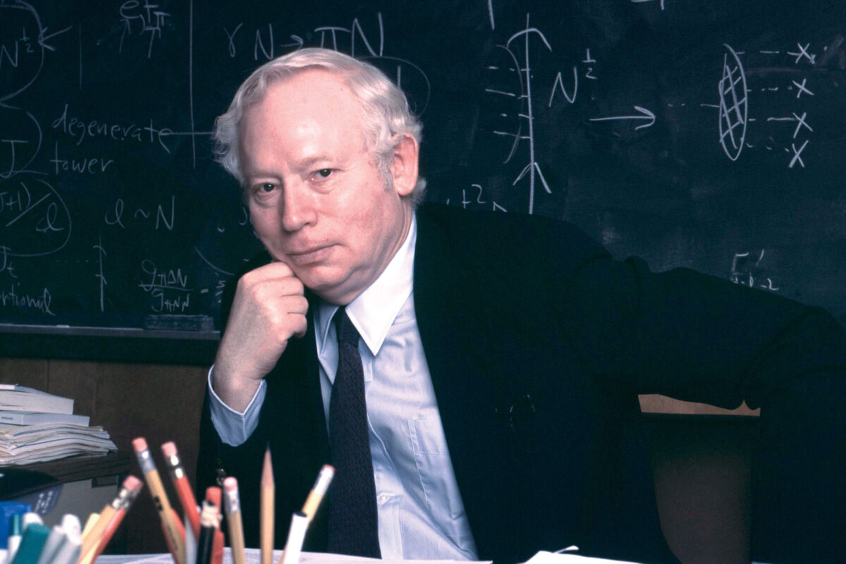 A man in a suit rests his chin on his hand while seated at a desk with a chalkboard and equations behind him